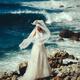 Woman in a white wedding dress standing on a rocky sea shore with a ve... (thumbnail)