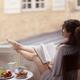 Woman reading a book with her legs on an open window frame and a plate... (thumbnail)
