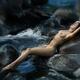 Woman lying naked on dark river rocks in rushing water stream Dramatic... (thumbnail)