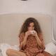 Pretty young woman in bohemian dress sitting on a white sofa sipping f... (thumbnail)