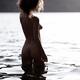Nude woman with curly hair standing in reflective water of a lake at s... (thumbnail)