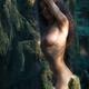 Beautiful nude woman standing leaning against a mossy tree in a dark s... (thumbnail)
