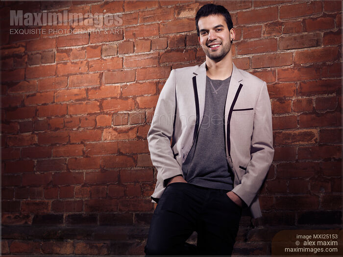 Stock photo of Artistic portrait of a young smiling man standing at brick wall Buy commercial use license at MaximImages
