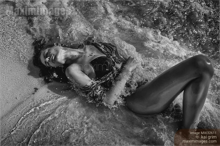 Stock photo of Beautiful sexy woman in swimsuit lying in sea water splashing waves hitting her body Dynamic black and white Buy commercial use license at MaximImages