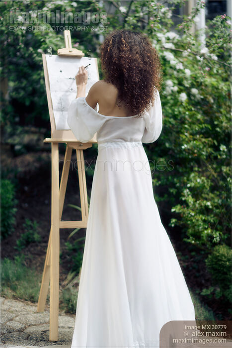 Stock photo of Beautiful woman artist with an easel painting outdoors in a garden in sheer white gown Buy commercial use license at MaximImages