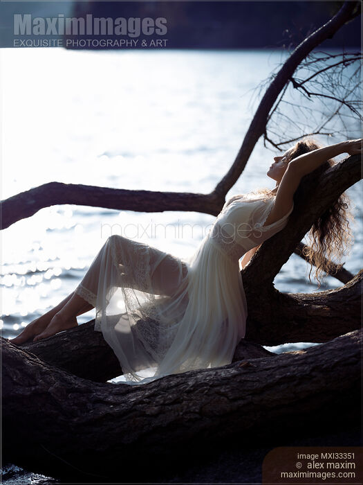 Beautiful woman in sheer white lace dress reclining relaxed against a tree by the lake in the nature Stock photo of Beautiful woman in sheer white lace dress reclining relaxed against a tree by the lake in the nature Buy commercial use license at MaximImages