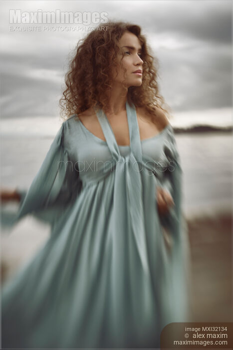 Stock photo of Beautiful young woman in a blue dress standing on the ocean shore with a thoughtful expressive look Buy commercial use license at MaximImages