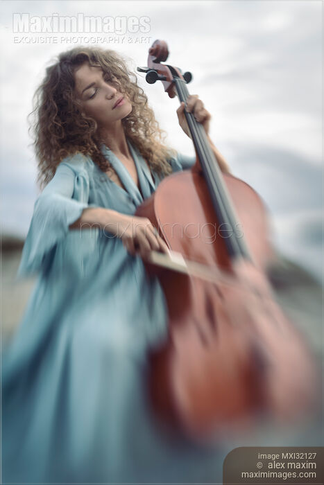 Stock photo of Beautiful young woman in a blue vintage dress passionately playing cello in sunrise nature scenery Buy commercial use license at MaximImages
