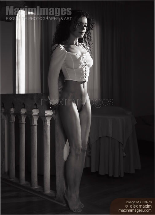 Stock photo of Black and white art nude portrait of a beautiful woman standing half-naked wearing only a top revealing her nudity in dark room Buy commercial use license at MaximImages