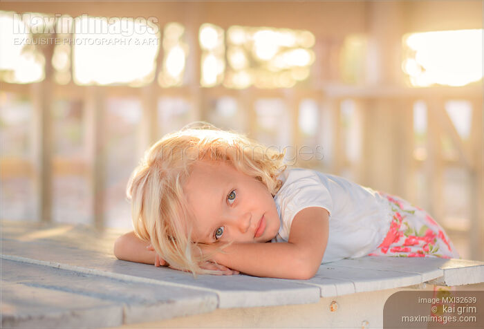 Stock photo of Cute little girl with blond hair and an expressive look in a sunlit gazebo at a tropical beach Buy commercial use license at MaximImages
