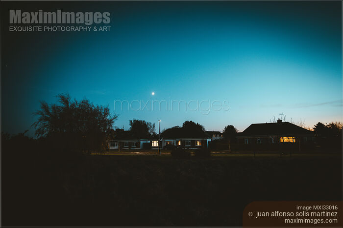 Stock photo of Dark residential houses with lit up windows on a street in dramatic night town scenery Buy commercial use license at MaximImages