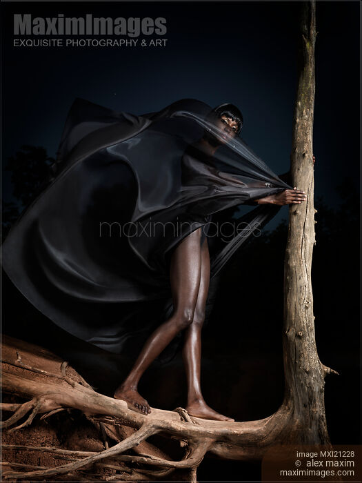Stock photo of Dramatic fine art portrait of black african woman standing barefoot on dead tree roots in dark night Buy commercial use license at MaximImages
