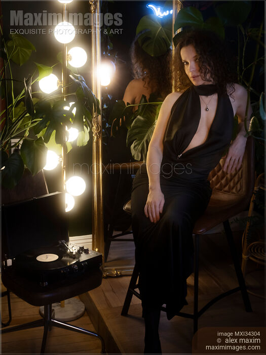 Stock photo of Elegant sexy woman in classy black silk dress sitting on a chair by a vinyl record player Buy commercial use license at MaximImages