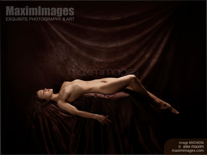 Fine art nude photograph of a beautiful woman lying naked posing in artist studio on dark background Stock photo of Fine art nude photograph of a beautiful woman lying naked posing in artist studio on dark background Buy commercial use license at MaximImages