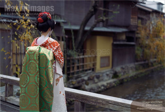 Stock photo of Maiko young Geisha apprentice in a red kimono with long obi standing on a bridge Buy commercial use license at MaximImages