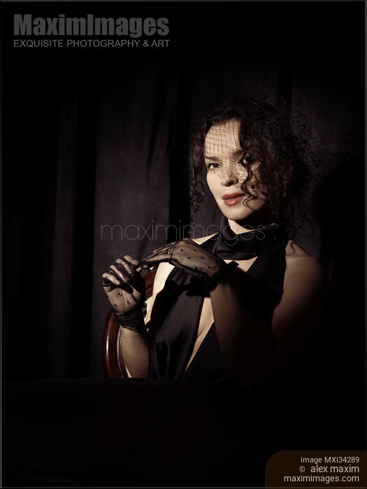 Stock photo of Portrait of beautiful mysterious femme fatale lady dressed in black wearing veiled hat taking off her gloves Buy commercial use license at MaximImages