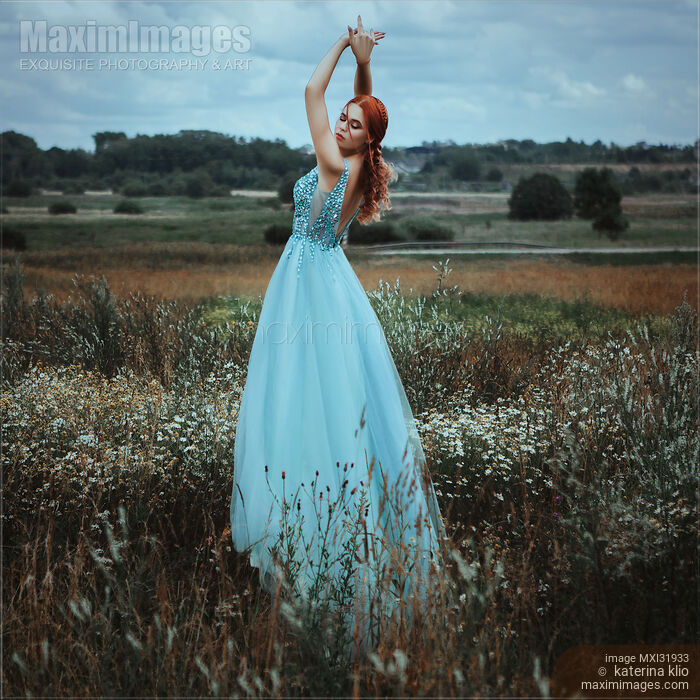 Stock photo of Red-haired woman in a long blue dress standing gracefully in a field of wild flowers Buy commercial use license at MaximImages