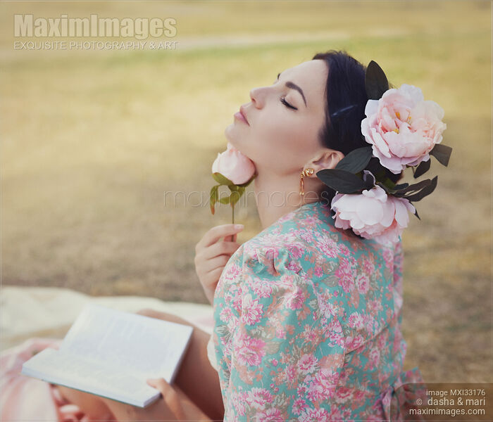 Stock photo of Romantic sensual portrait of a beautiful woman with pink flowers in her hair reading a book outdoors in a park Buy commercial use license at MaximImages