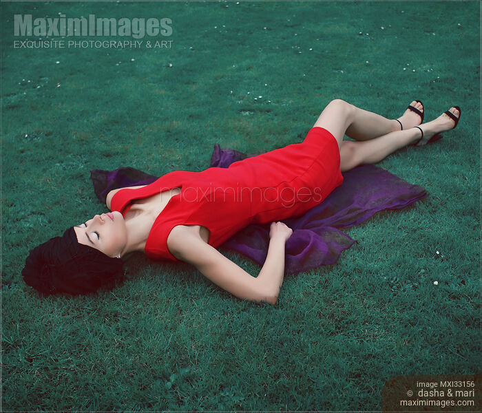Stock photo of Surreal fashion portrait of extravagant woman in bright red dress lying on blue green grass Buy commercial use license at MaximImages