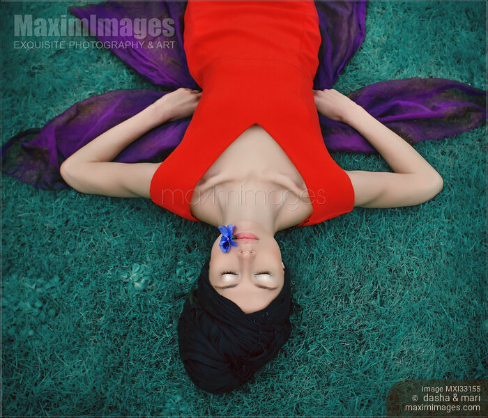 Stock photo of Surreal portrait of an extravagant lady in red dress lying on blue grass with a flower in her mouth Buy commercial use license at MaximImages