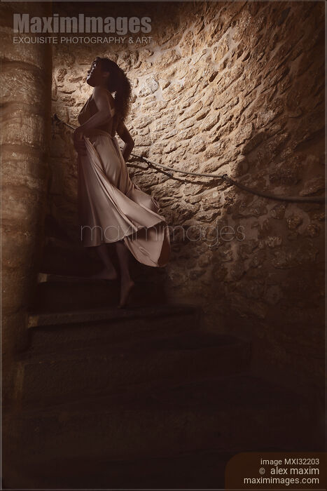 Stock photo of Woman in a dress walking cautiously up a dark shadowy spiral staircase in a dungeon of a French house Buy commercial use license at MaximImages