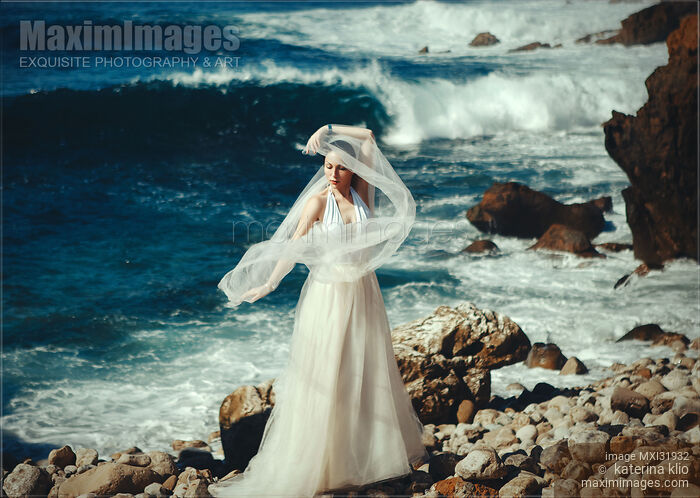 Stock photo of Woman in a white wedding dress standing on a rocky sea shore with a veil in the wind Buy commercial use license at MaximImages