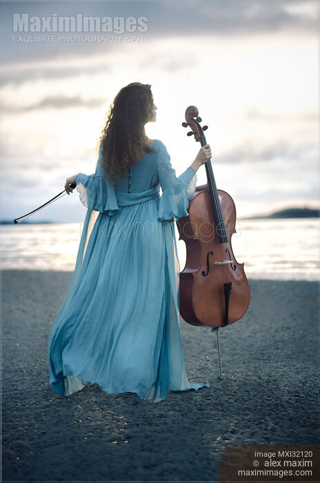 Stock photo of Woman in blue dress standing on a sea shore holding a cello Rear view of her back in dramatic sunrise Buy commercial use license at MaximImages