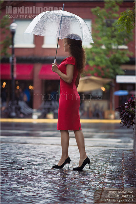 Stock photo of Woman in elegant red dress waiting for someone on a city street with an umbrella under the rain Buy commercial use license at MaximImages