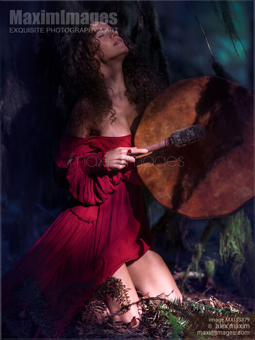 Stock photo of Woman in red dress playing shamanic drum in tranquil dark forest nature scenery Buy commercial use license at MaximImages