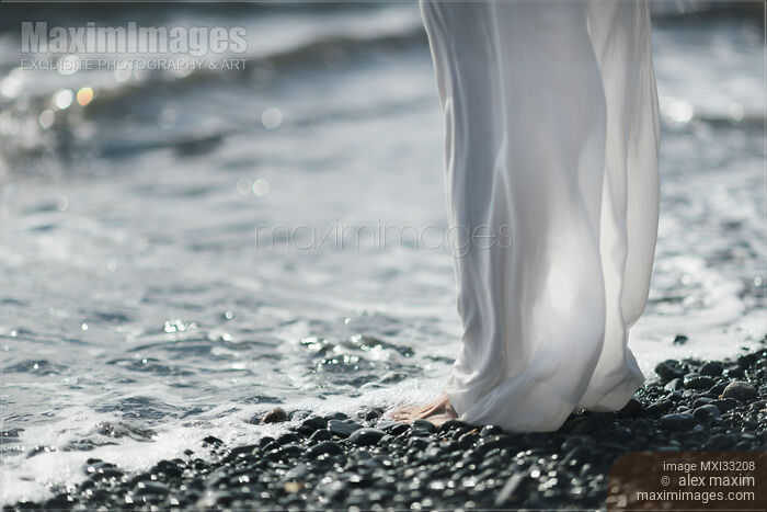 Stock photo of Woman in white summer pants standing barefoot on a pebbled ocean beach shore, cool sea water washing over her bare feet Buy commercial use license at MaximImages