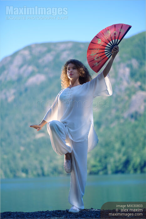 Stock photo of Woman practicing Tai Chi crane stance with a fan at the mountain lake shore in the morning Buy commercial use license at MaximImages