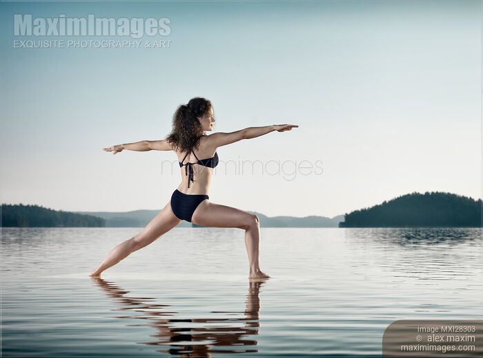 Stock photo of Woman practicing yoga on the water doing Warrior pose over blue sky Buy commercial use license at MaximImages