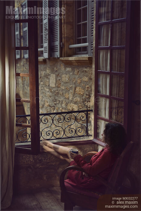 Woman sitting thoughtfully in a chair by an open windows in France holding a cup of coffee in her hand Stock photo of Woman sitting thoughtfully in a chair by an open windows in France holding a cup of coffee in her hand Buy commercial use license at MaximImages