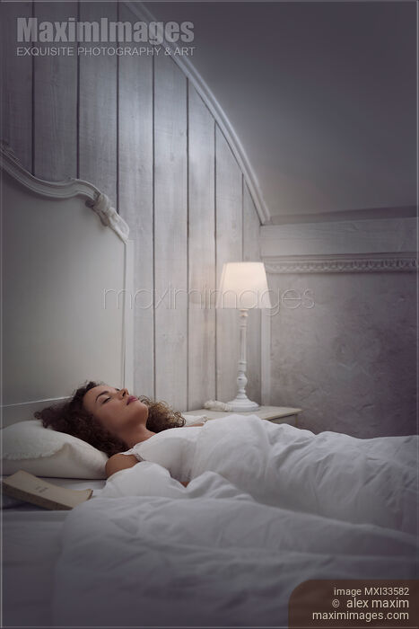 Stock photo of Woman sleeping in bed alone in white vintage room with a book and a lamp beside her Buy commercial use license at MaximImages