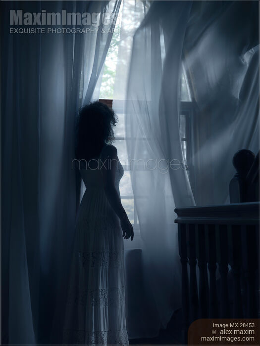 Stock photo of Woman standing at window in dark room in dim sunlight Buy commercial use license at MaximImages