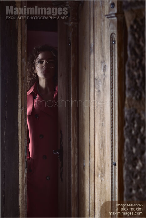 Stock photo of Woman standing behind a slightly opened entrance door of a house looking out at a night visitor Buy commercial use license at MaximImages