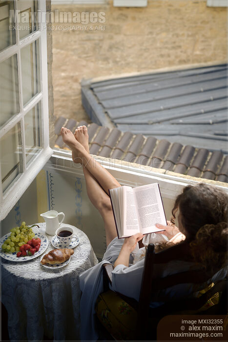 Stock photo of Young French woman reading book by an open window in the morning light Buy commercial use license at MaximImages