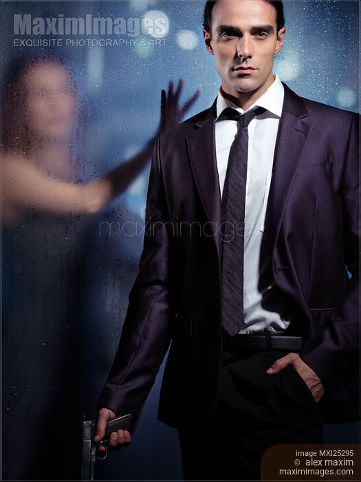 Stock photo of Young man in a suit with a gun in his hand and woman behind glass. Dramatic couple relationship portrait. Buy commercial use license at MaximImages