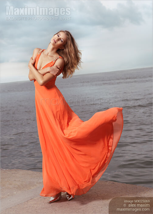 Stock photo of Young woman in orange dress flying in the wind at sea shore Buy commercial use license at MaximImages