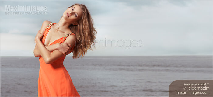 Stock photo of Young woman in orange dress standing on a shore Buy commercial use license at MaximImages