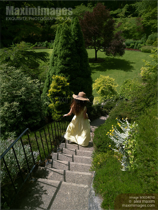 Stock photo of Young woman in summer dress and hat running down the stairs in a green garden Buy commercial use license at MaximImages