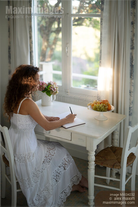 Stock photo of Young woman in white summer dress writing in a diary sitting at a table by a window in country house Buy commercial use license at MaximImages